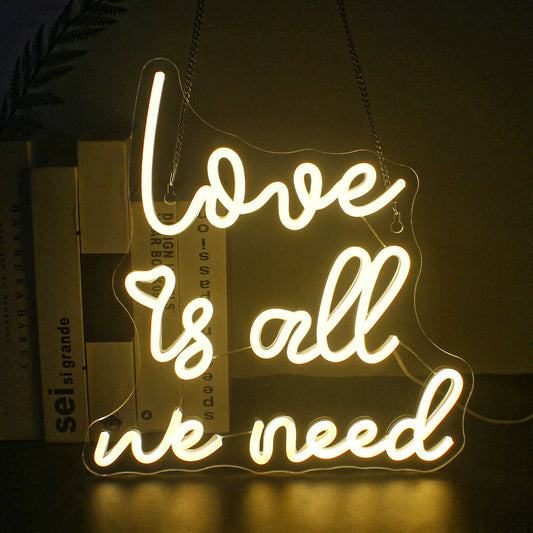 The image features a vibrant LED neon sign that reads "Love is all we need" in a flowing, cursive font.