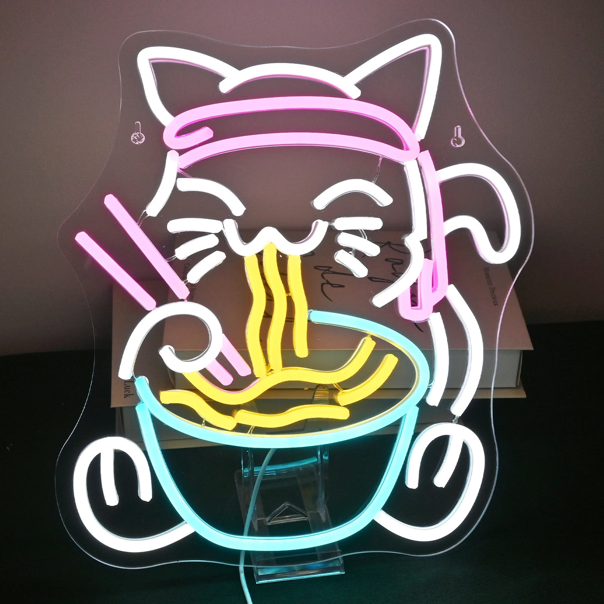 Bilden har en levande och färgglad LED-neonskylt som visar en nyckfull design av en katt som njuter av en skål med ramen.