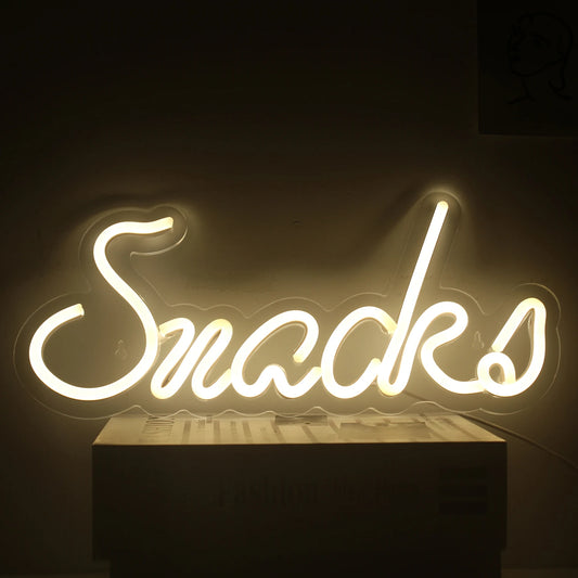 Bilden visar en levande LED-neonskylt i form av ordet "Snacks" i ett lekfullt, kursivt teckensnitt.