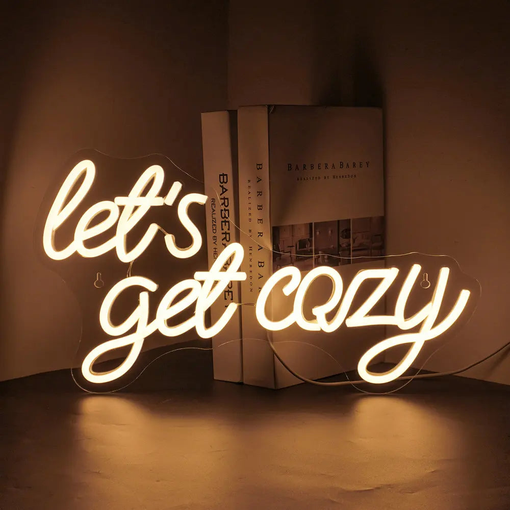 Bilden har en levande LED-neonskylt som visar frasen "Let's Get Cozy" i ett varmt, välkomnande manustypsnitt.