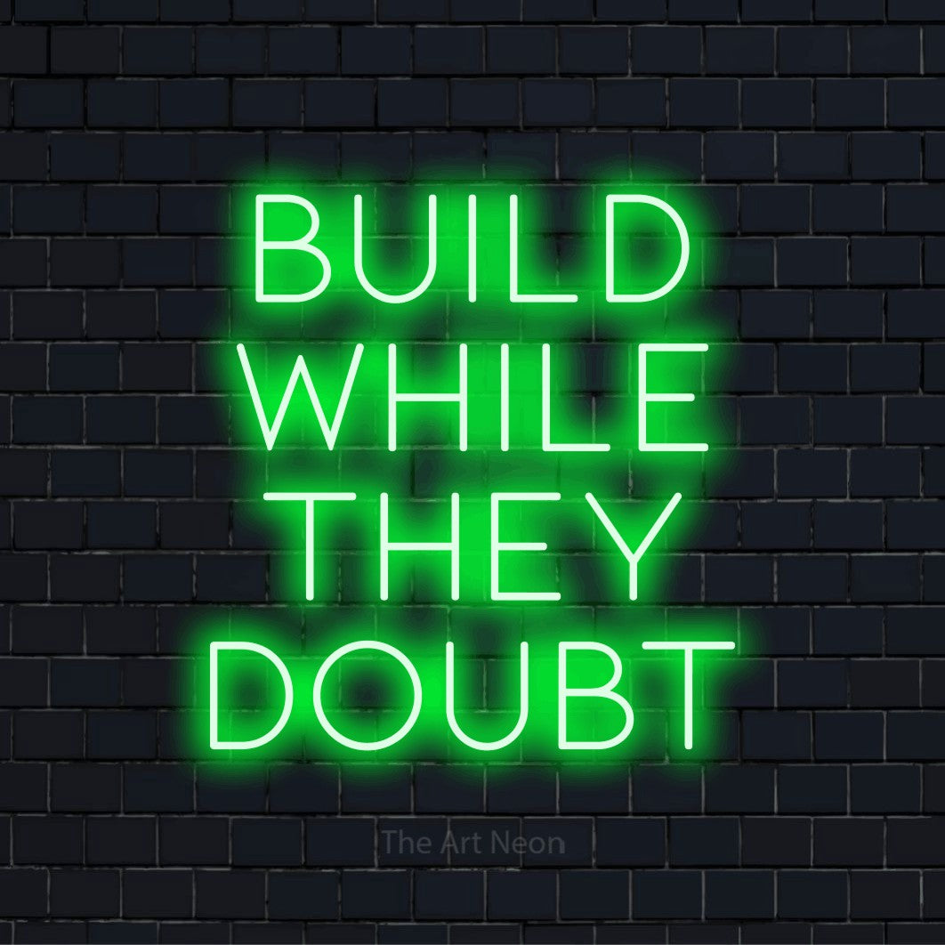 "Build While They Doubt" script-style LED neon sign on a black brick wall